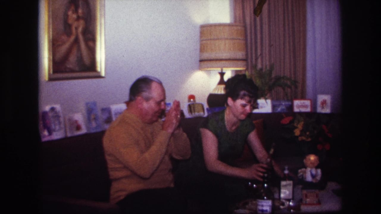 1967 Rockaway Beach home-movie frame showing a man in a mustard sweater and a woman on a couch near drinks and Christmas cards.