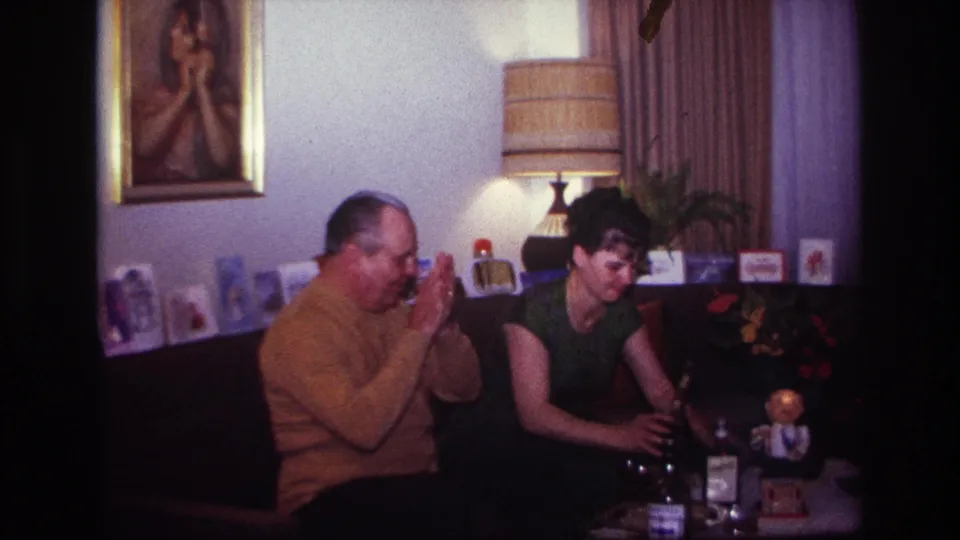 1967 Rockaway Beach home-movie frame showing a man in a mustard sweater and a woman on a couch near drinks and Christmas cards.