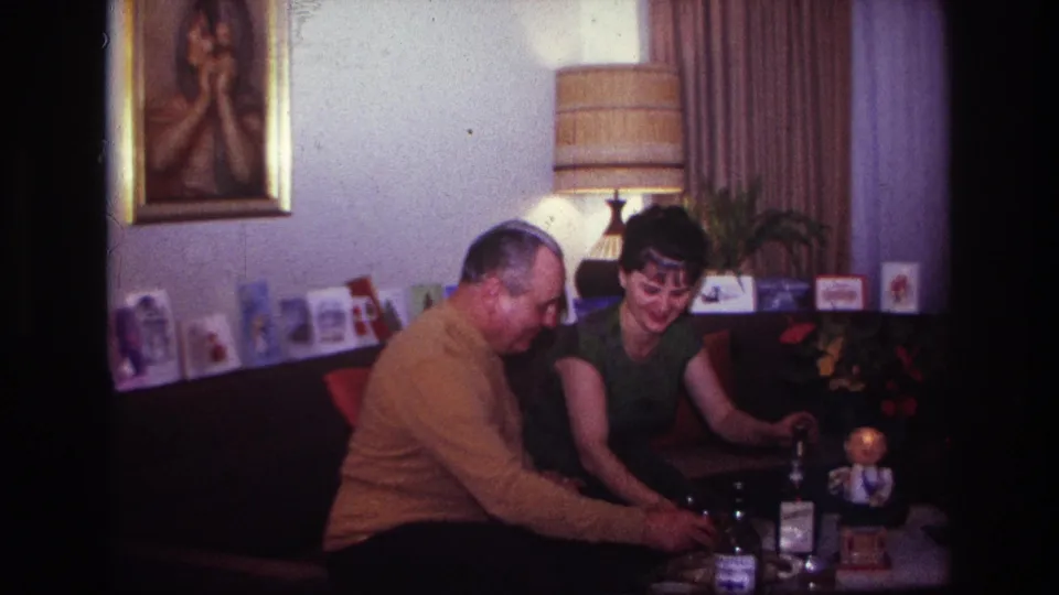 Rockaway Beach 1967 home-movie frame of two adults on a couch handling drinks beneath a lamp and row of Christmas cards.