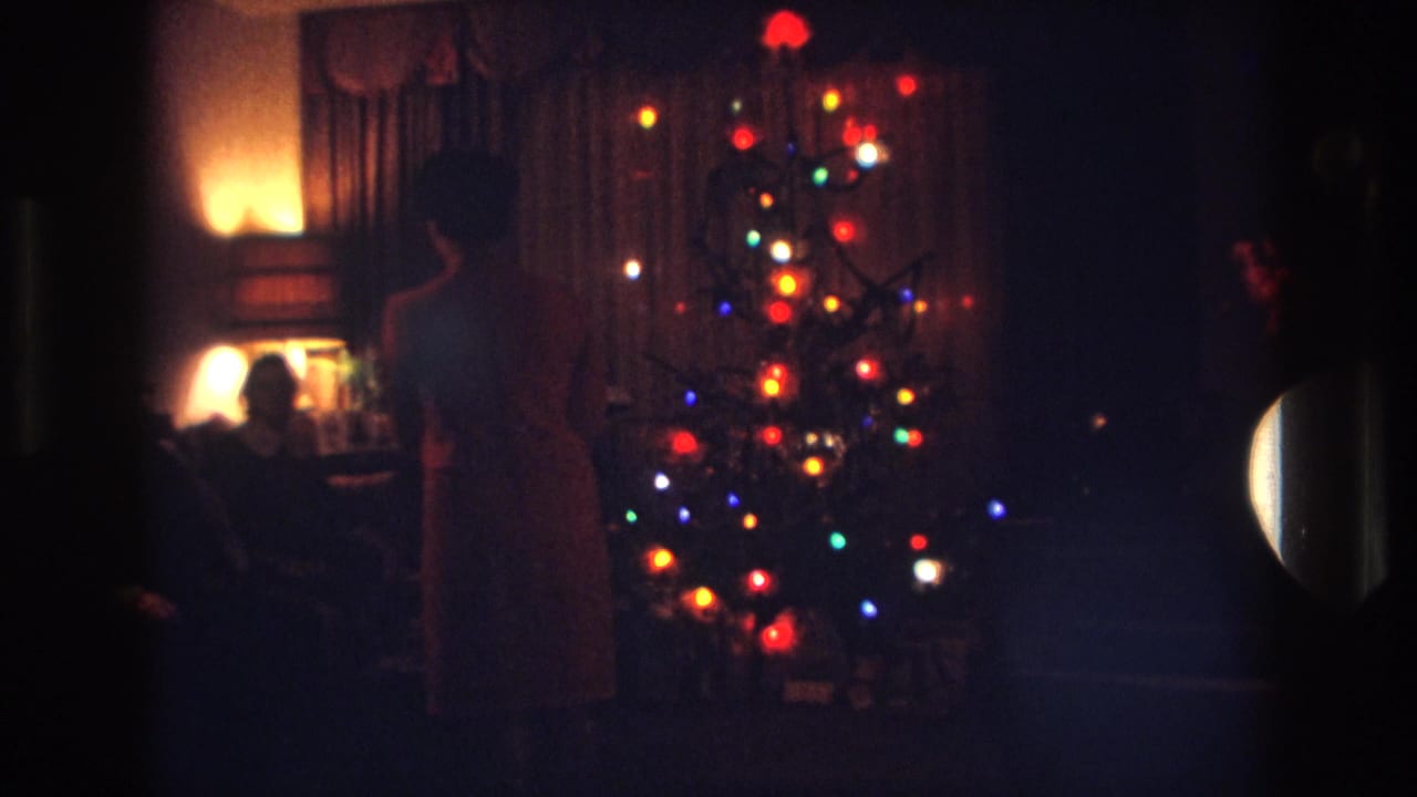 1967 Rockaway Beach home-movie frame showing a dim holiday living room, colored Christmas tree lights, and family members in shadow.