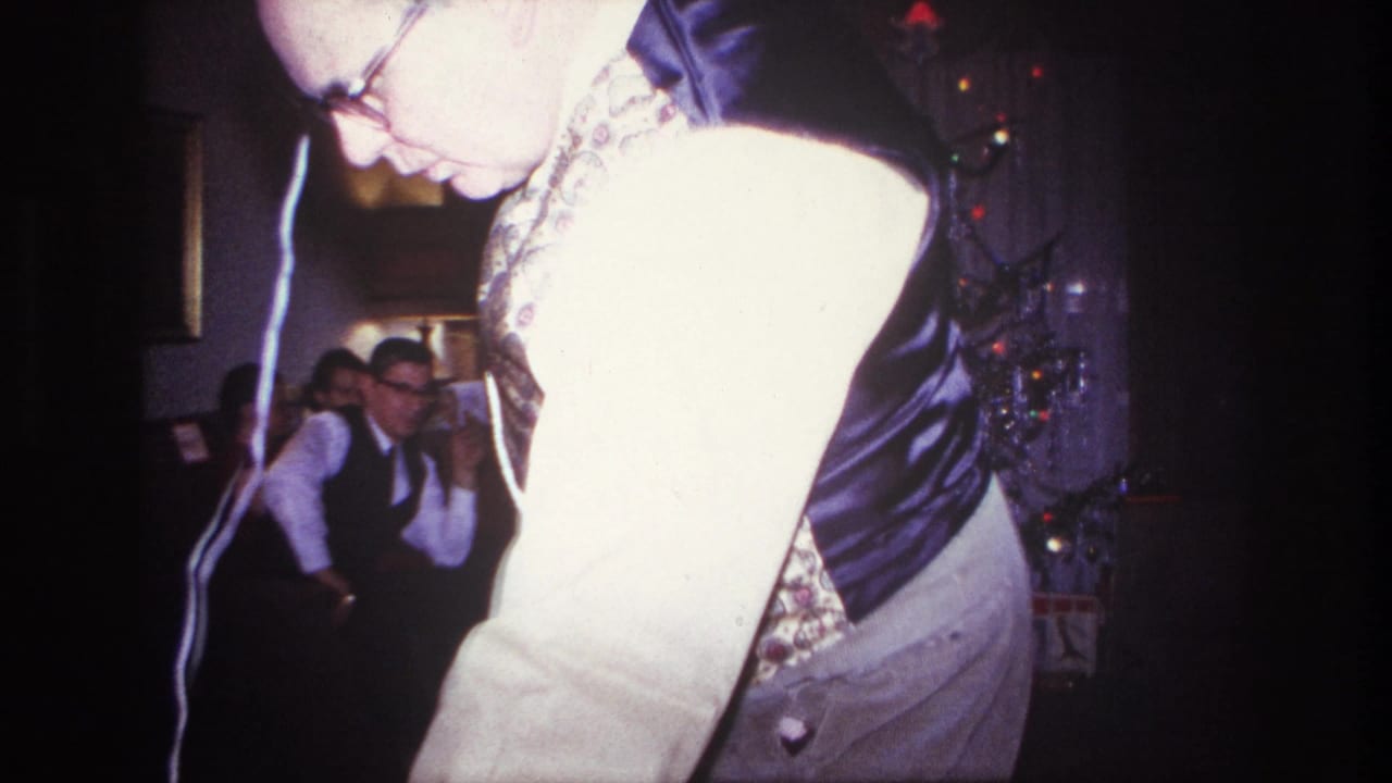 Man with glasses and patterned vest turns near the camera beside a Christmas tree.
