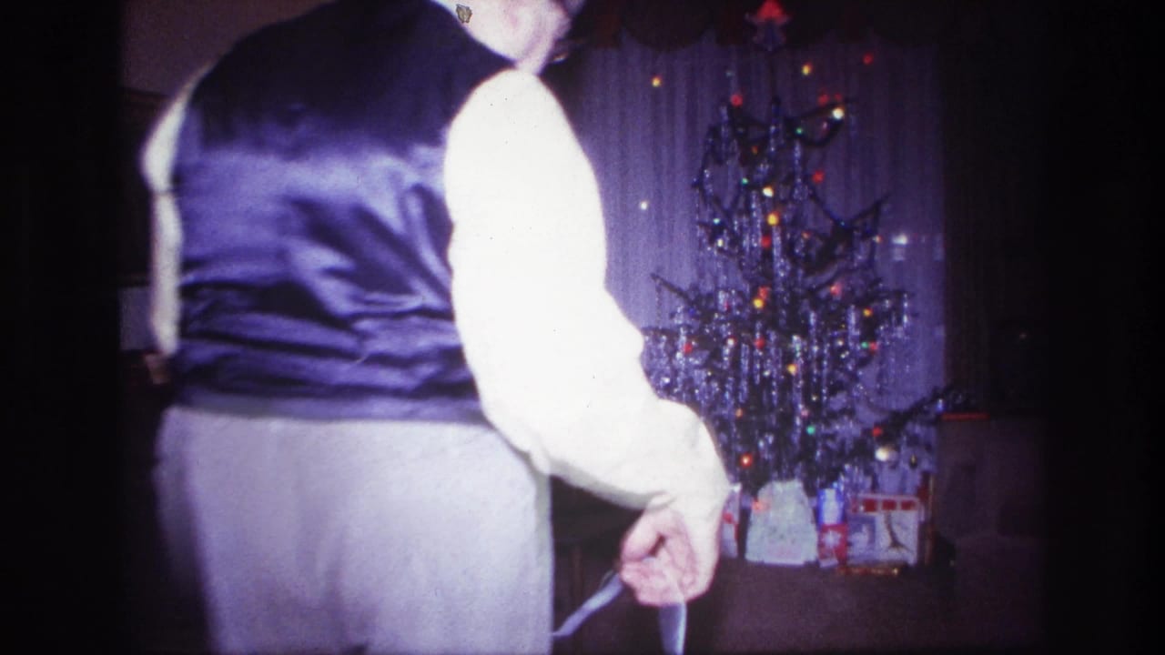 1967 Rockaway Beach home-movie frame with a man in formal clothing near a tinsel Christmas tree in a dim living room.