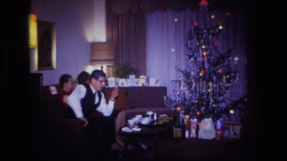 Adults gather on a sofa in a Rockaway Beach living room beside a tinsel Christmas tree, with gifts and tableware visible.