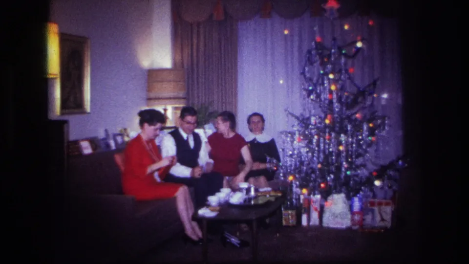 Adults in formal clothing sit and talk beside a Christmas tree in a private Rockaway Beach living room in 1967.