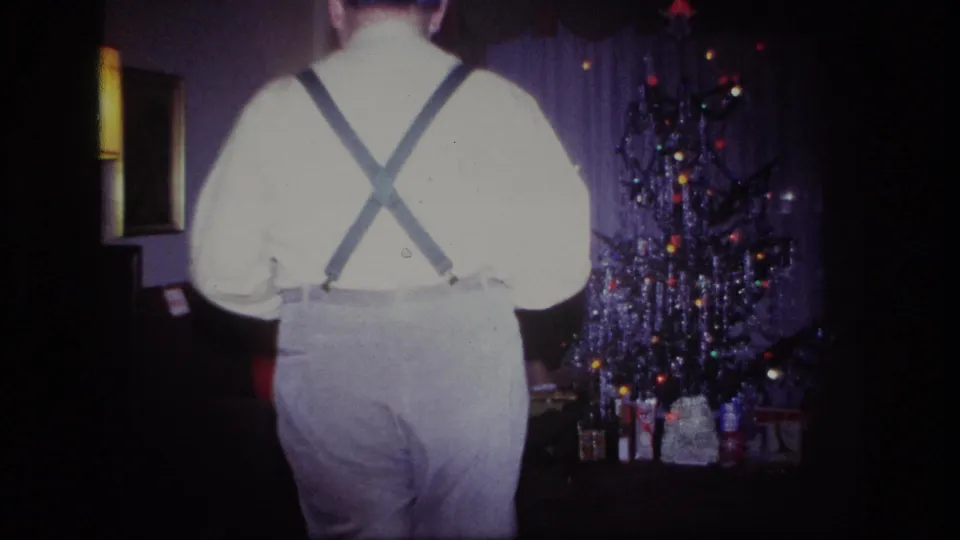 A man in suspenders crosses in front of the camera in a 1967 home-movie view of a Christmas living room.