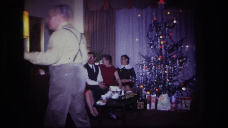 A man in suspenders moves through the foreground while adults sit by a tinsel Christmas tree in Rockaway Beach, 1967.