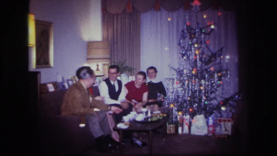 1967 holiday home-movie frame with adults seated on a sofa in a Rockaway Beach living room beside a silver tinsel tree.