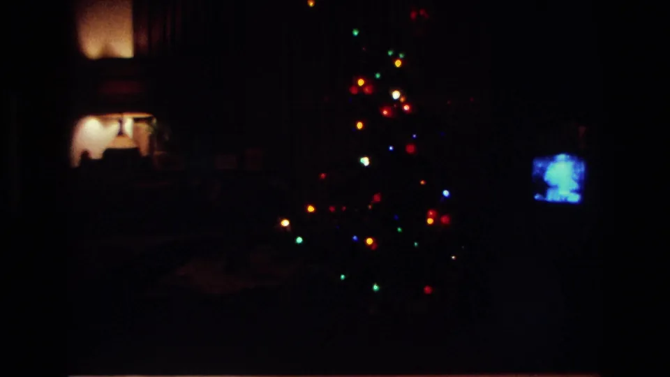 Dim 1967 home-movie view of a private living room with Christmas lights glowing and a small television screen at right.