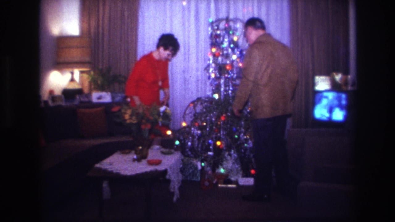 1967 Rockaway Beach home-movie frame showing a woman in red and a man in a brown jacket decorating a silver tinsel Christmas tree.