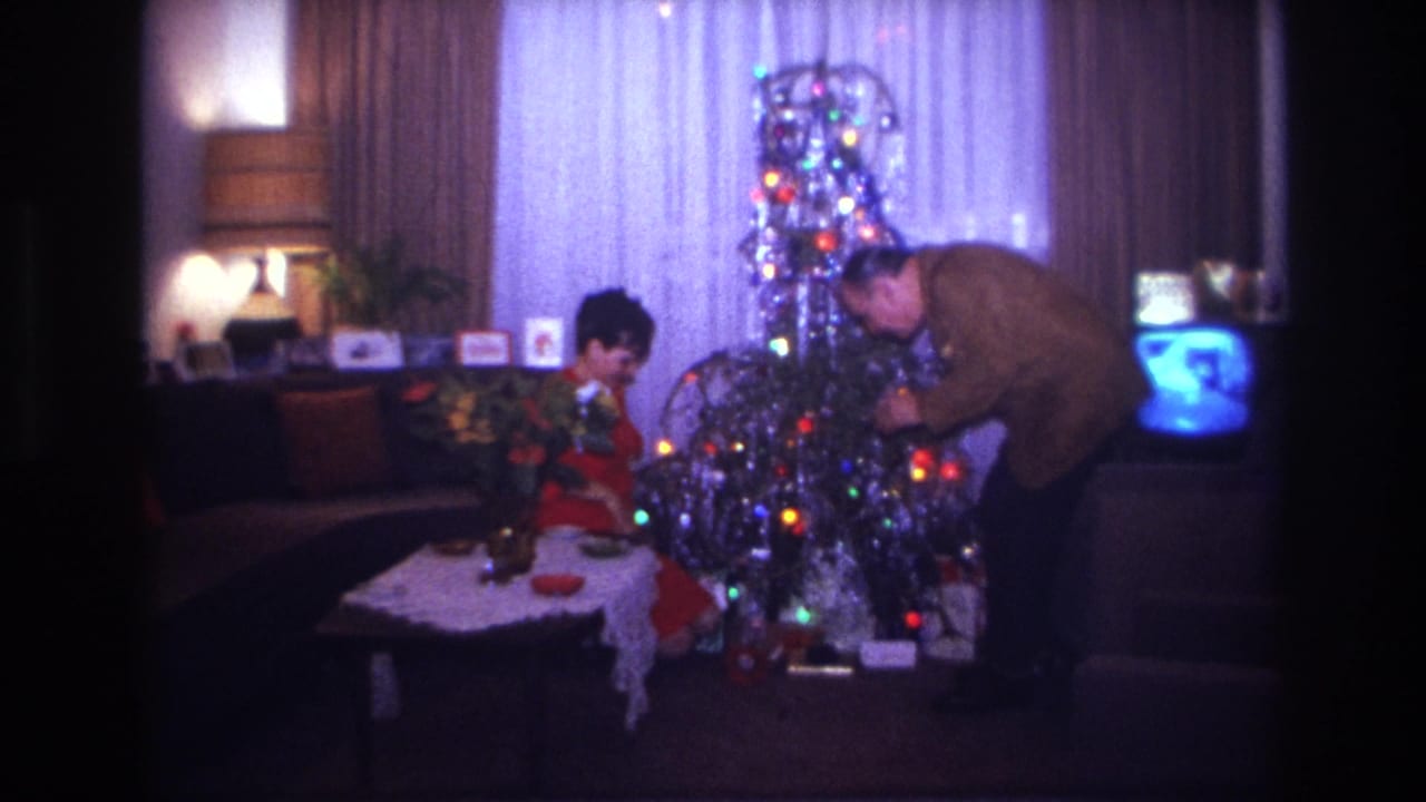 A seated woman and a standing man decorate a lit silver Christmas tree beside a glowing television.