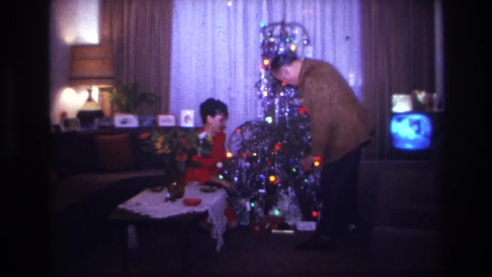 Home-movie frame from 1967 Rockaway Beach showing Christmas tree decorating in a dim private living room with a television at right.