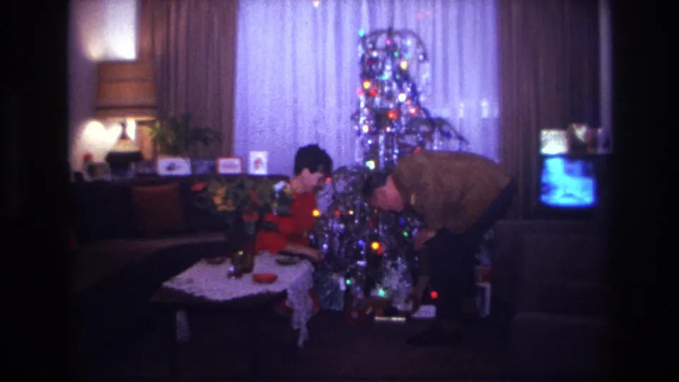 1967 Rockaway Beach Christmas decorating frame with a man bending near a lit silver tinsel tree and a woman seated beside it.