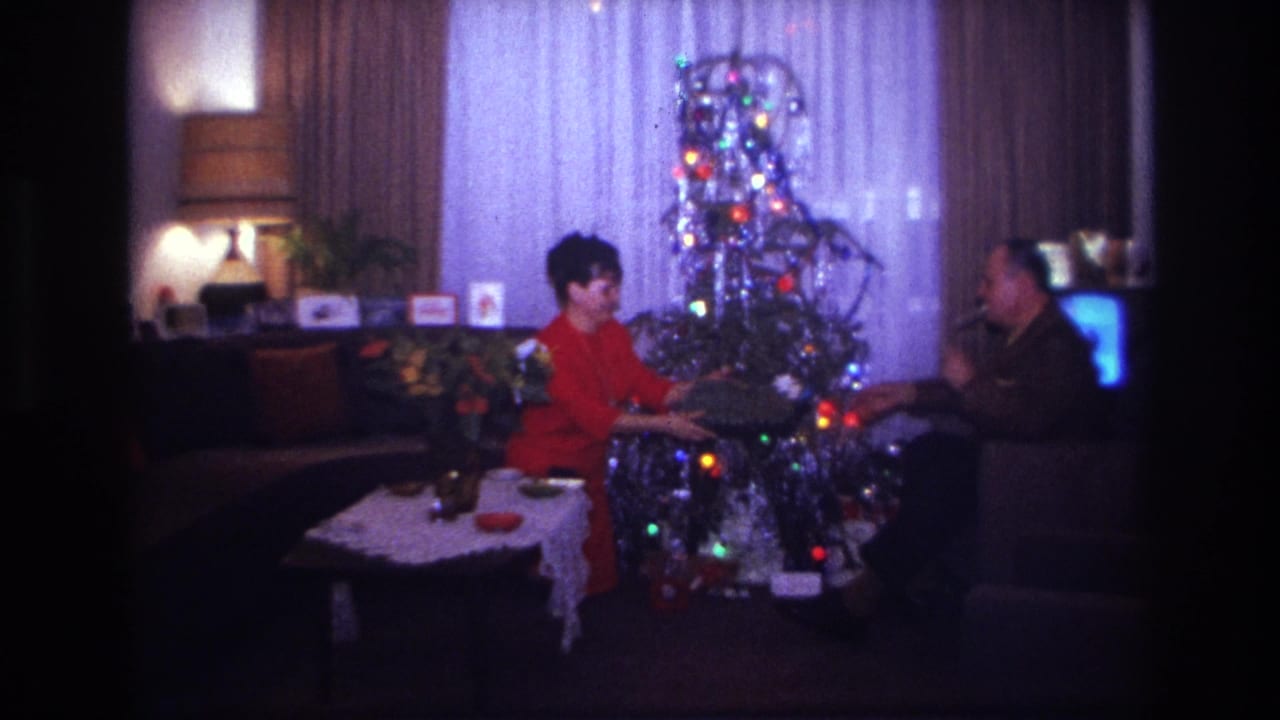In a 1967 Rockaway Beach home movie frame, two adults sit by a silver Christmas tree during a quiet gift exchange.