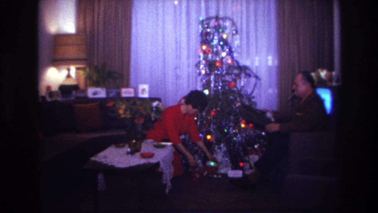 Woman leans toward gifts beside the tree while a man watches from a chair.