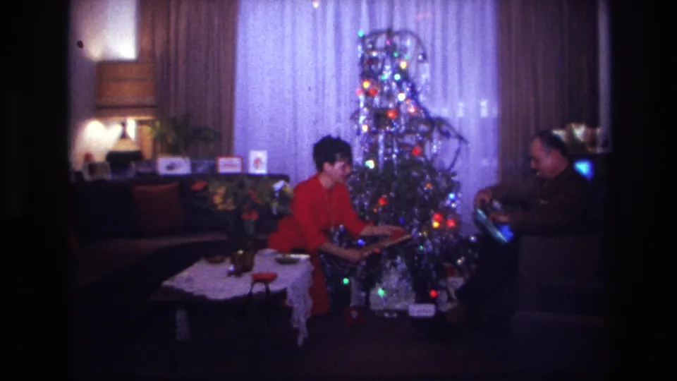 The woman presents or opens a package near the silver tree while the seated man looks on in the Rockaway Beach interior.