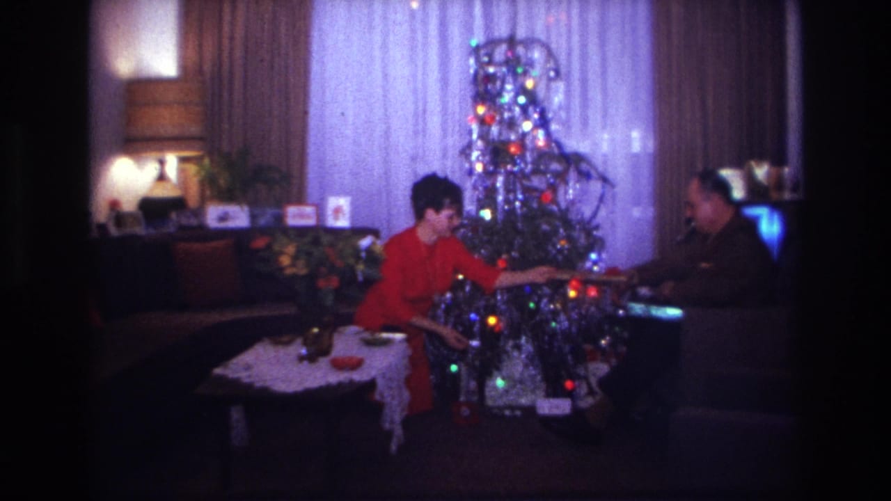 1967 Rockaway Beach home-movie frame showing a woman in red adjusting a lit Christmas tree as a seated man watches in a living room.
