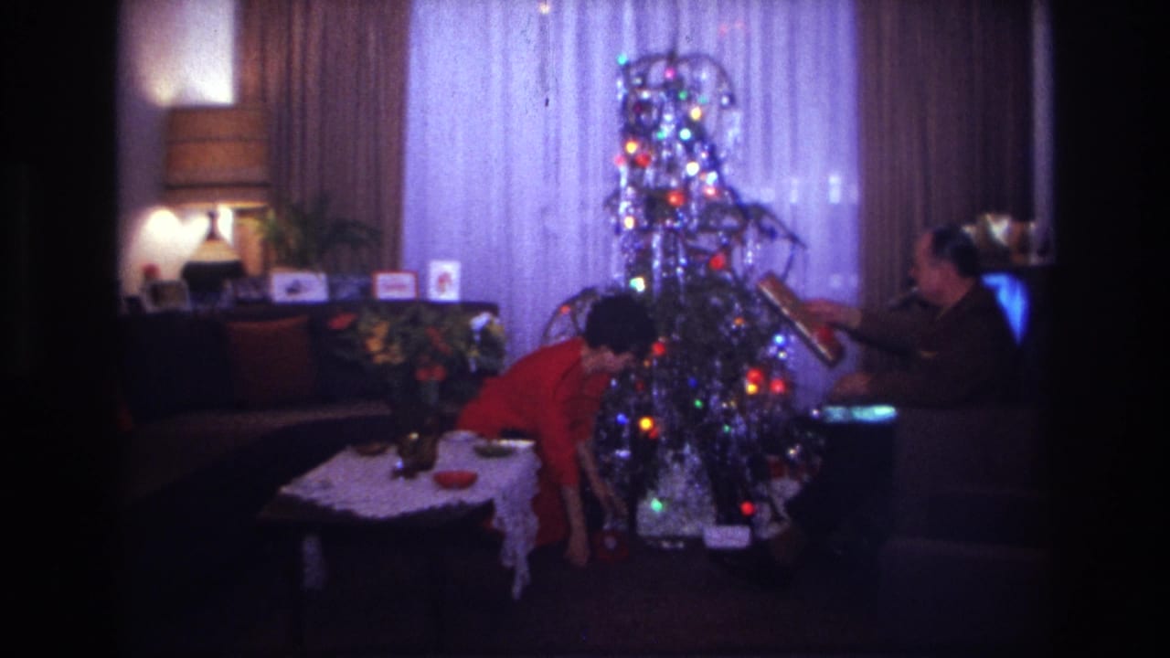 Woman bends near the lit Christmas tree as a seated man reaches toward the decorations.