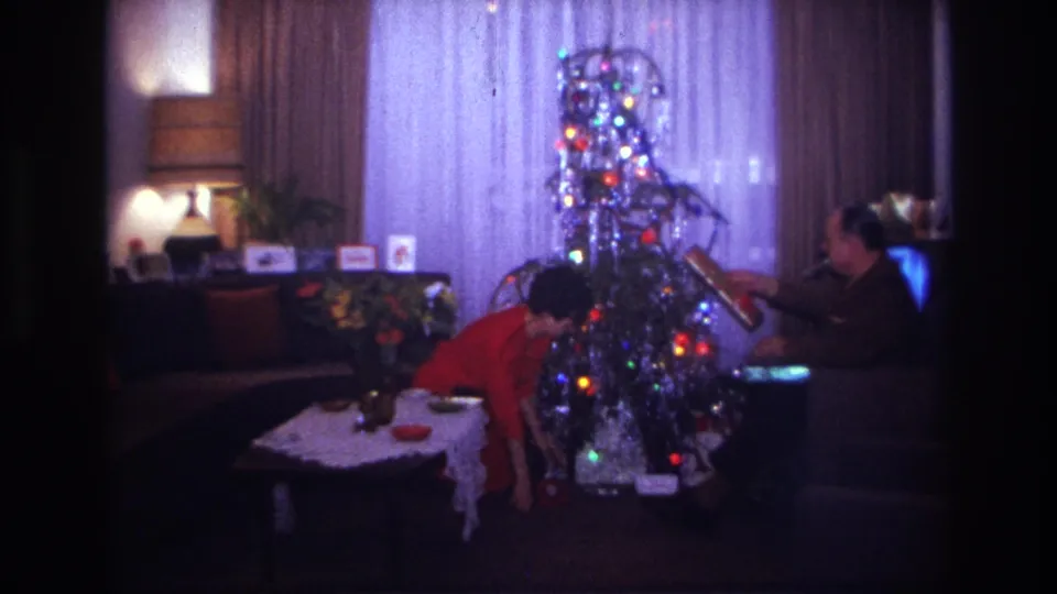 1967 Rockaway Beach home-movie frame with a woman bending at the Christmas tree while a man sits beside the lights and tinsel.