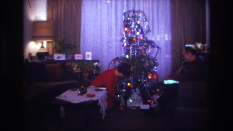 1967 Rockaway Beach home-movie frame showing a woman arranging items near the base of a lit Christmas tree as a man sits to the side.