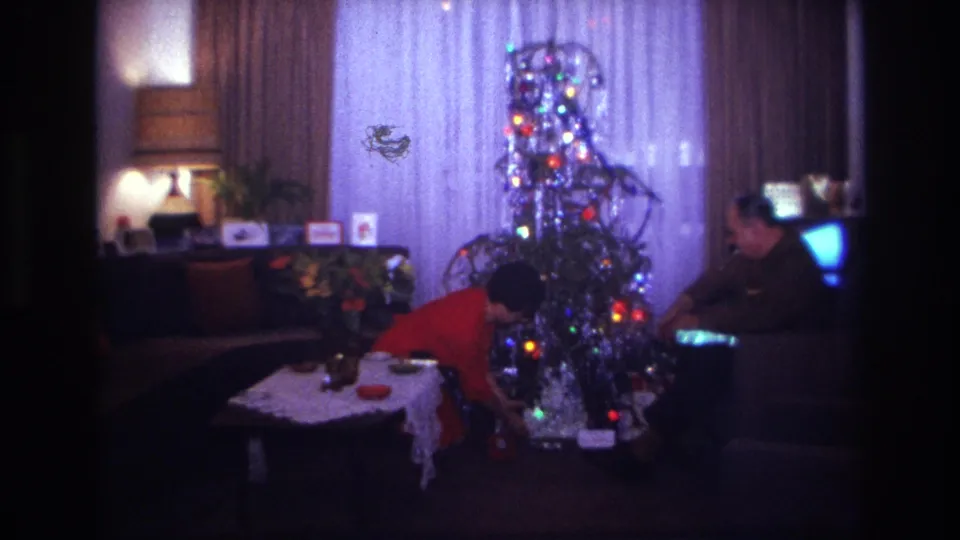 1967 Rockaway Beach home-movie frame of a woman bending beside a tinsel-covered Christmas tree, with a seated man and glowing television nearby.