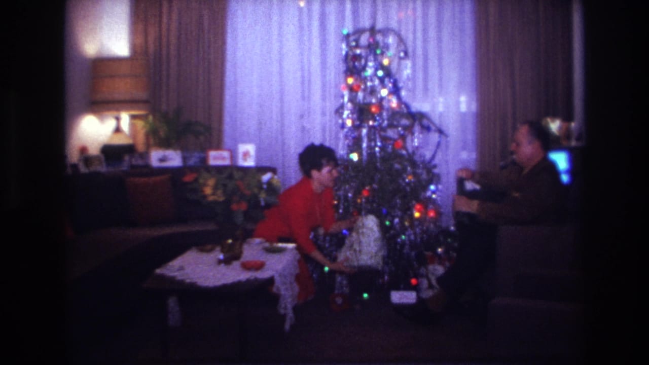 1967 Rockaway Beach home-movie frame showing a woman in red offering a wrapped gift beside a silver tinsel Christmas tree.
