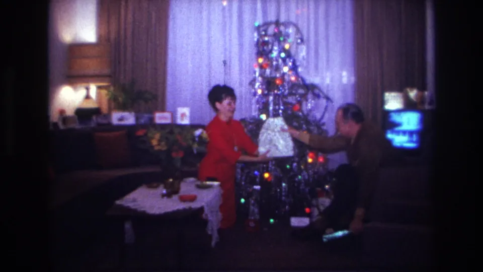 1967 Rockaway Beach holiday interior showing a woman and man exchanging a large wrapped gift beside a lit tinsel tree.