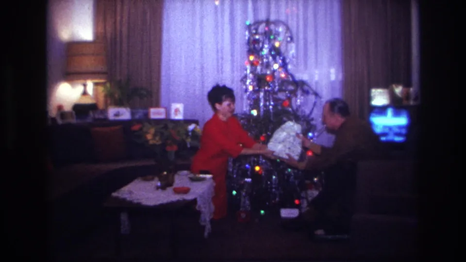 1967 Rockaway Beach home-movie frame of a man receiving a wrapped gift from a woman in a decorated living room.