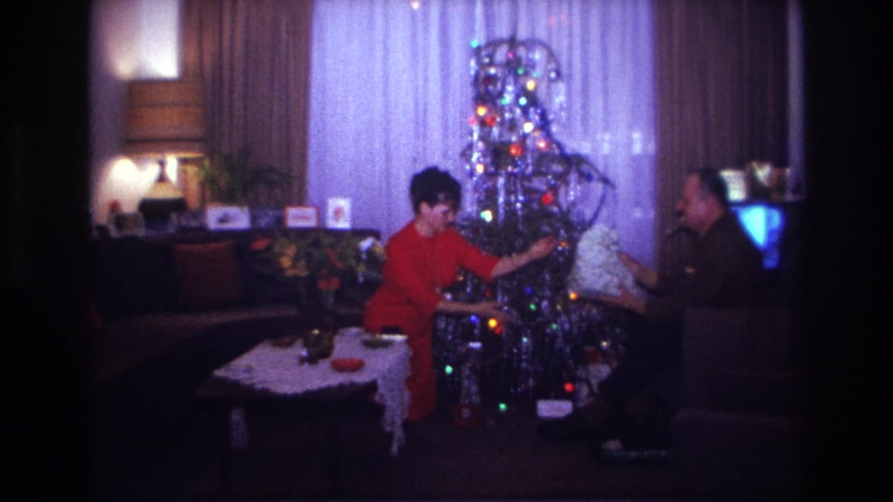 1967 Rockaway Beach home-movie frame showing two adults by a lit silver tinsel Christmas tree as a gift is passed across the room.