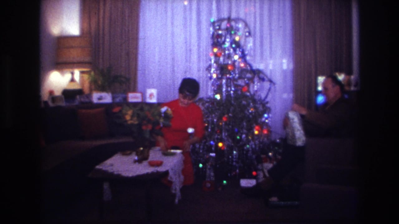 A woman looks down at a gift while a man sits near the Christmas tree holding wrapping paper.