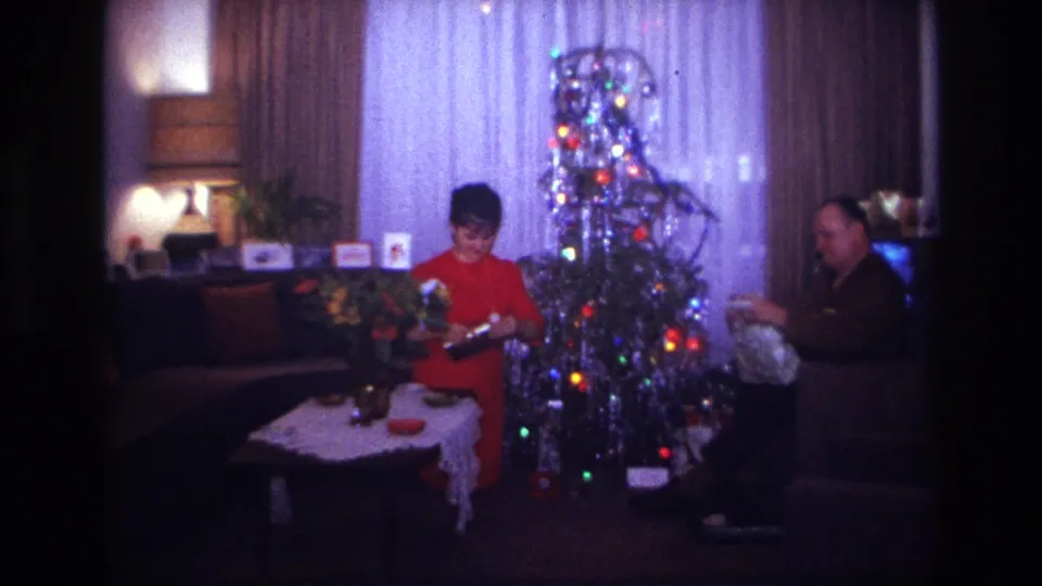 1967 Rockaway Beach home-movie frame of two adults by a lit silver tinsel Christmas tree after opening presents in the living room.