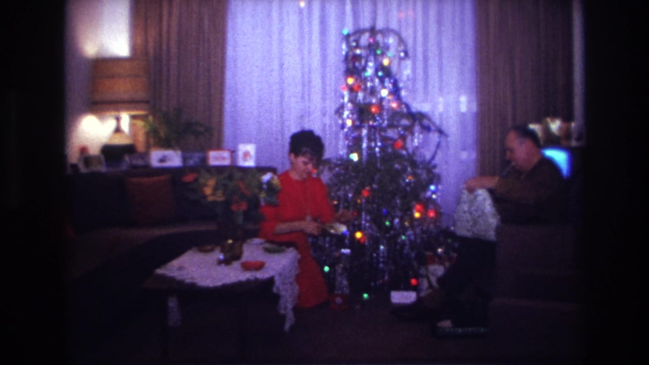 1967 Rockaway Beach home-movie frame showing two adults beside an aluminum Christmas tree in a private living room.