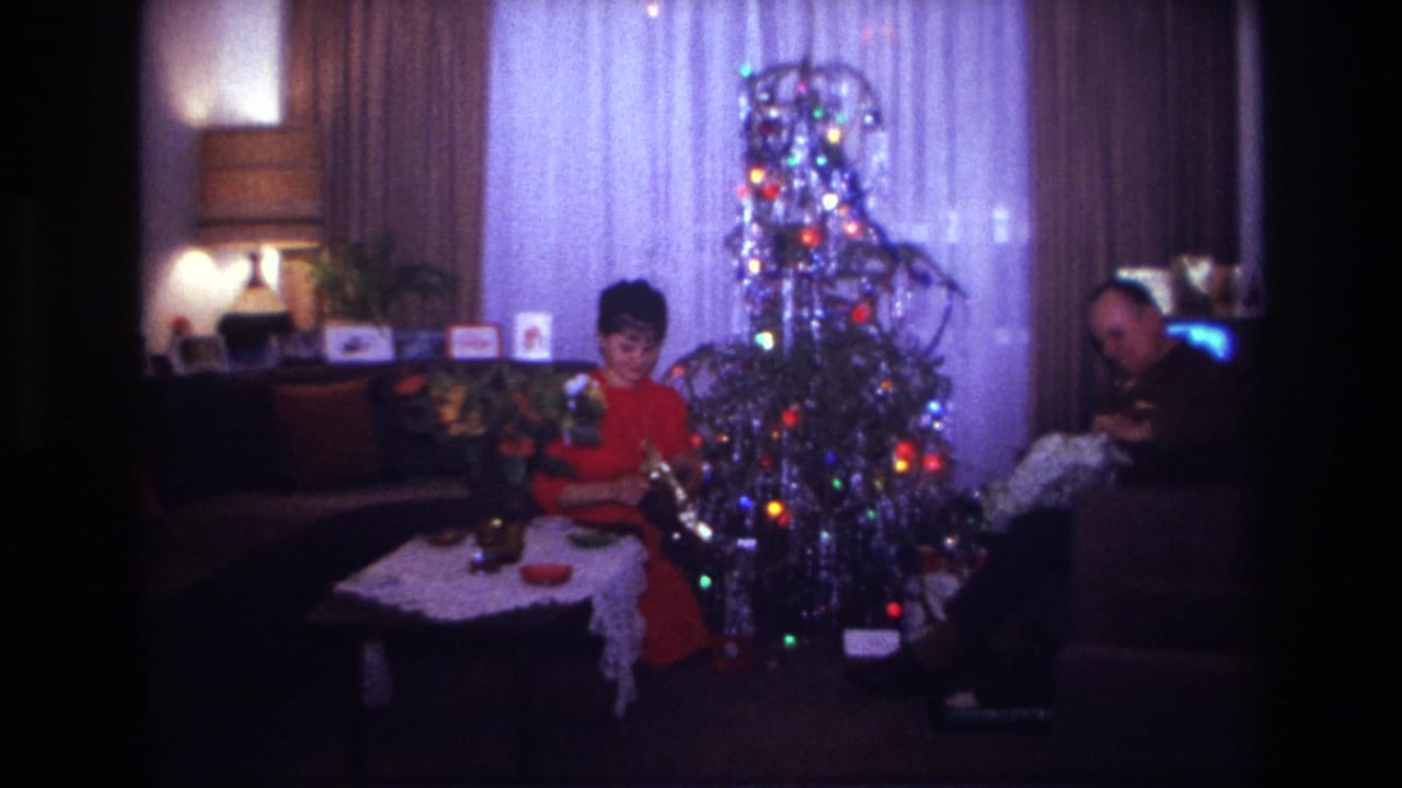 1967 Rockaway Beach home-movie frame showing adults opening Christmas presents beside a decorated tree in a private living room.