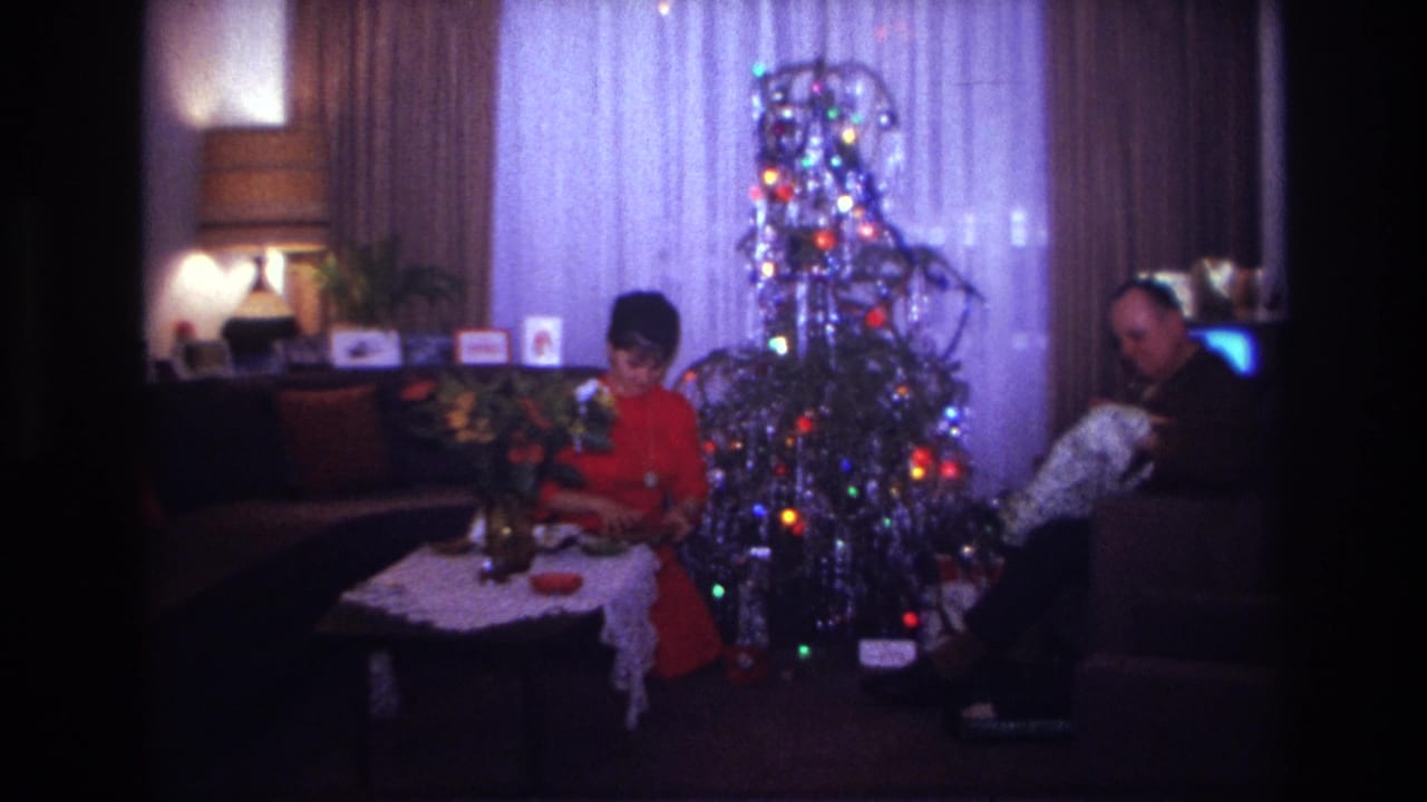 Woman handles a gift beside a lit Christmas tree while a man sits at right.