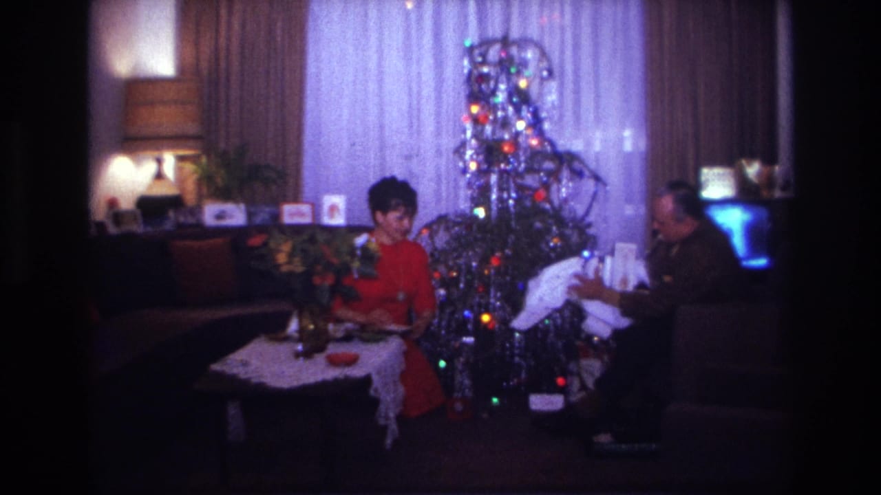 1967 Rockaway Beach home-movie frame showing two adults opening Christmas gifts beside a lit silver tinsel tree in a private living room.