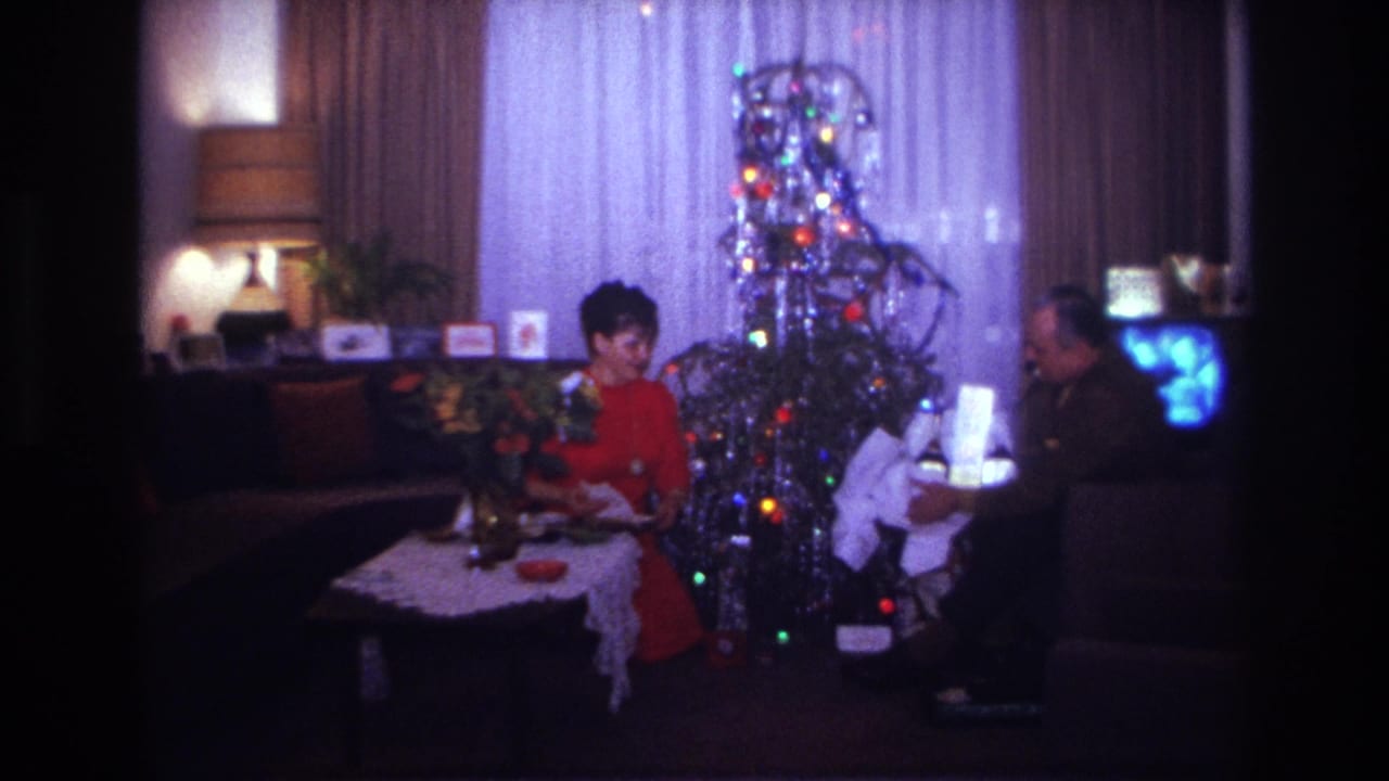 A woman in red and a seated man handle gifts near a lit tinsel Christmas tree.