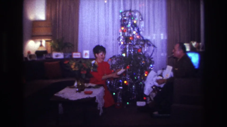 In the Rockaway Beach Christmas home movie, the woman examines a gift item while the man sits near the illuminated silver tinsel tree.