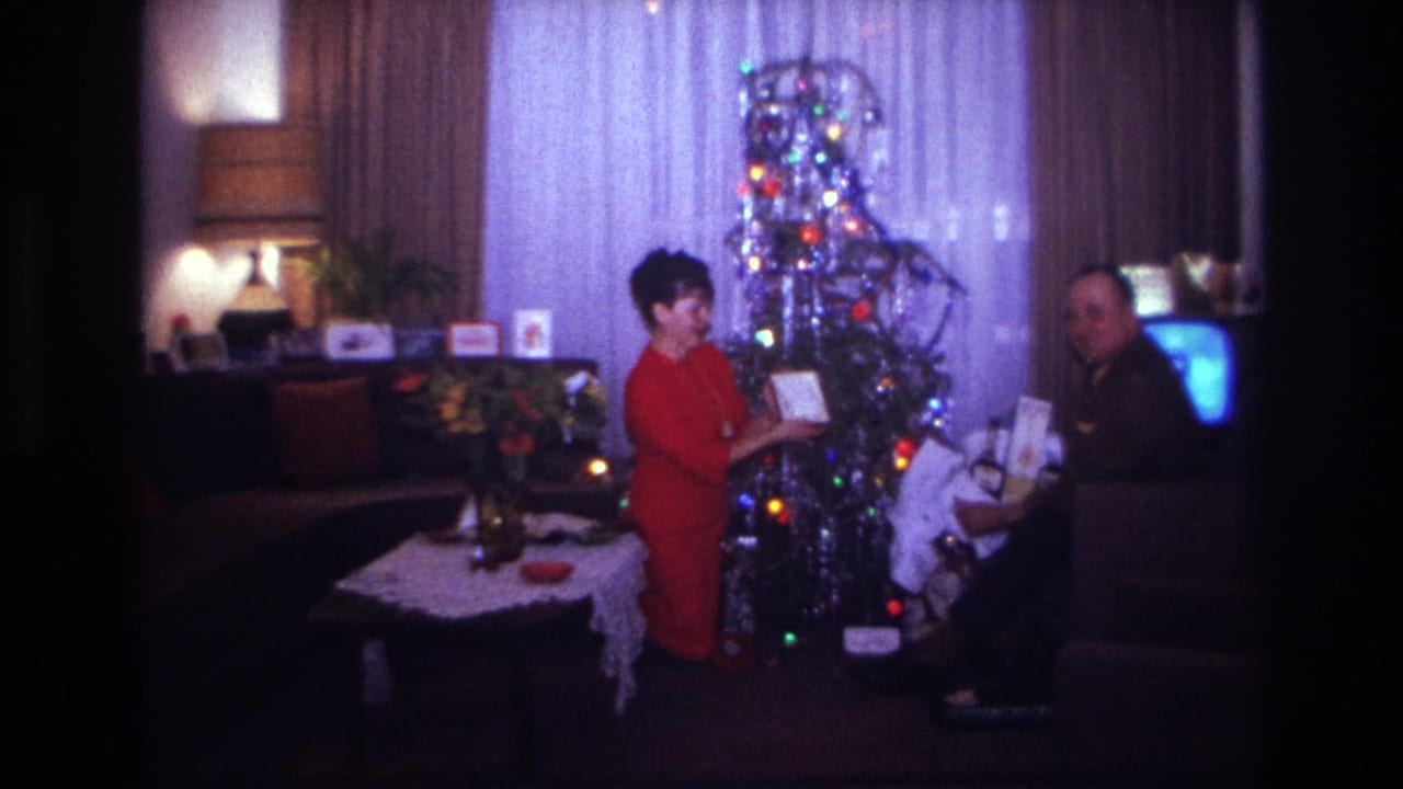 In a 1967 Rockaway Beach home movie frame, a woman in red stands by a decorated Christmas tree while a seated man holds a gift.