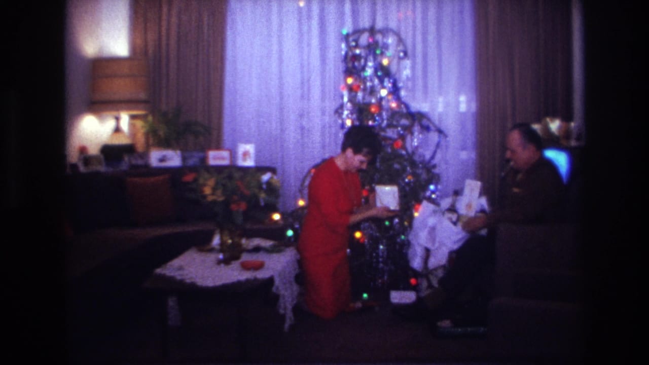A woman bends toward a seated man while handing or receiving a gift near the Christmas tree.