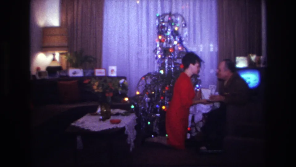 Two adults share a Christmas gift moment in a Rockaway Beach living room, with a lit tree, sofa, framed photos, and television visible.