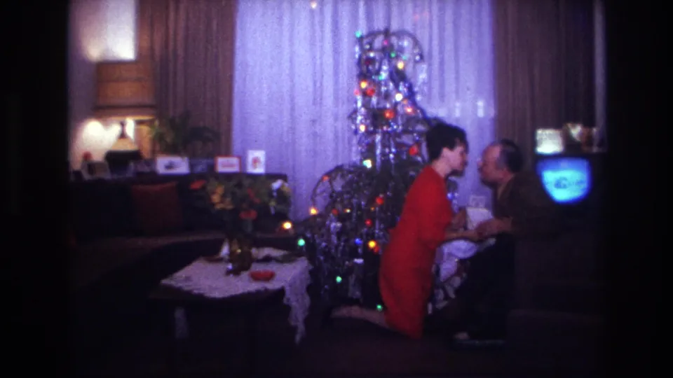 1967 Rockaway Beach home-movie frame of two adults kissing near a tinsel Christmas tree and glowing television.