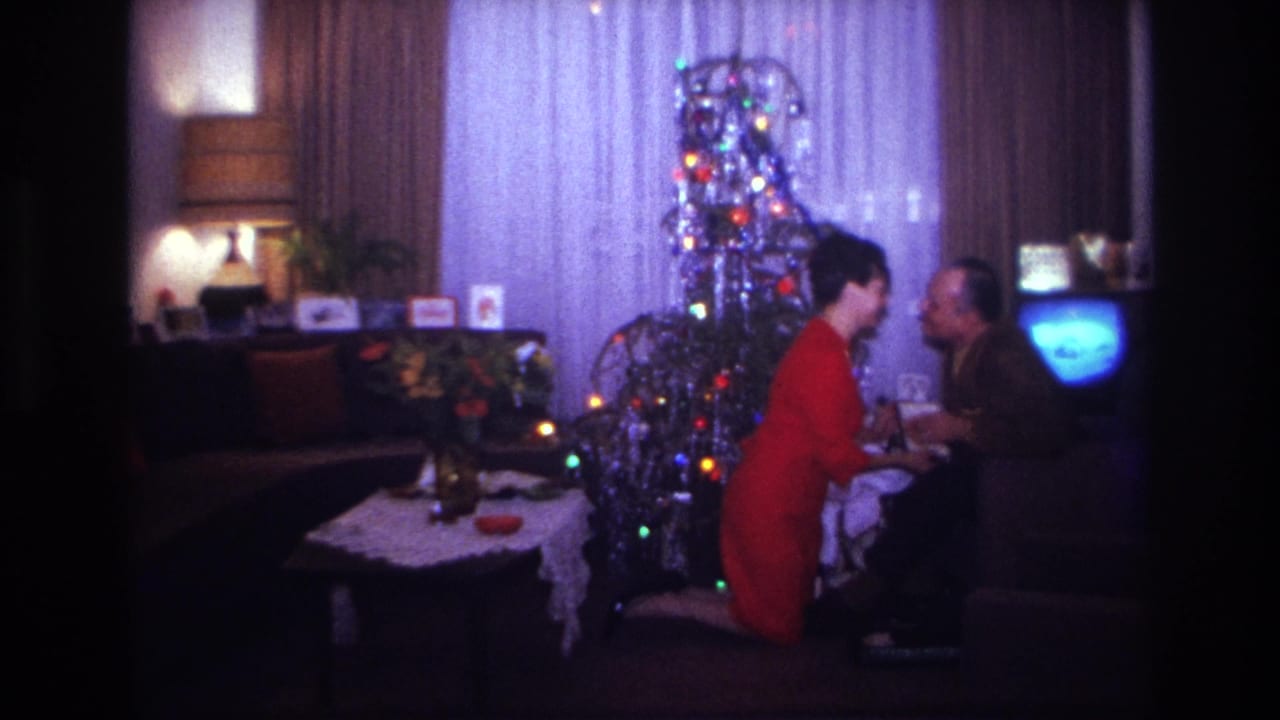 Couple leaning close by a Christmas tree and glowing television.