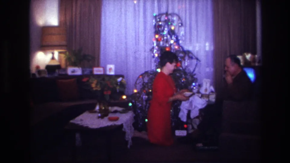 1967 home-movie frame showing the woman turning from the seated man near presents, a tinsel tree, flowers, and living-room furniture.