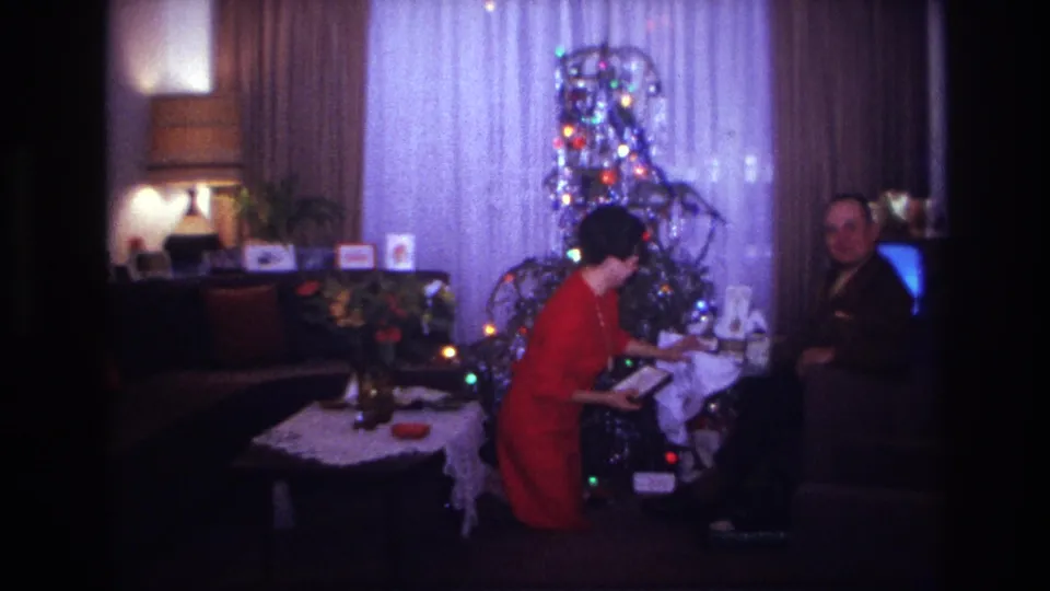 The woman bends near the presents while the man looks toward the camera beside the glowing television in the Christmas living room.