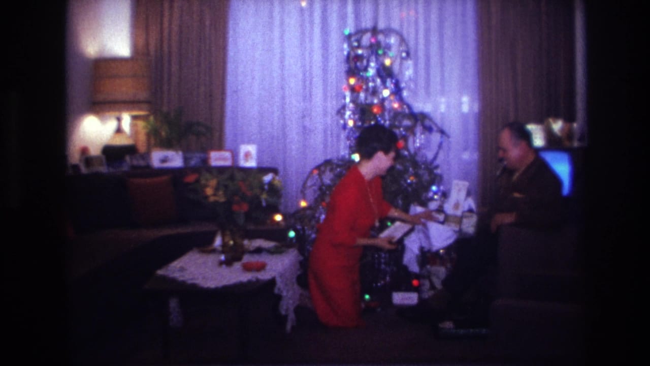 1967 Rockaway Beach home-movie frame showing a woman in red and a man exchanging gifts beside a lit tinsel Christmas tree.