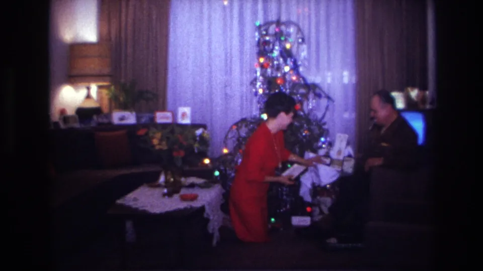1967 Rockaway Beach home-movie frame showing a woman in red and a man exchanging gifts beside a lit tinsel Christmas tree.