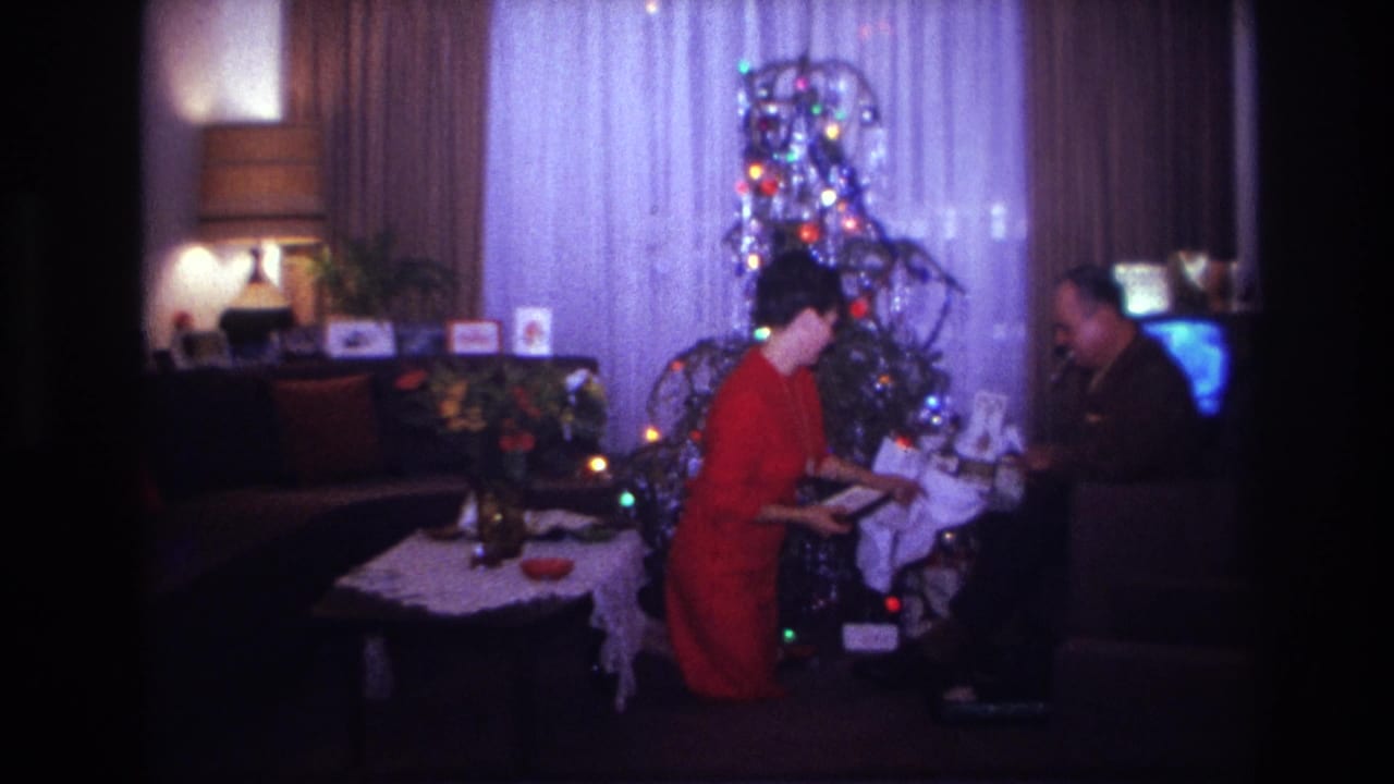 1967 Rockaway Beach interior frame with two adults exchanging Christmas gifts beside a lit tinsel tree and television.