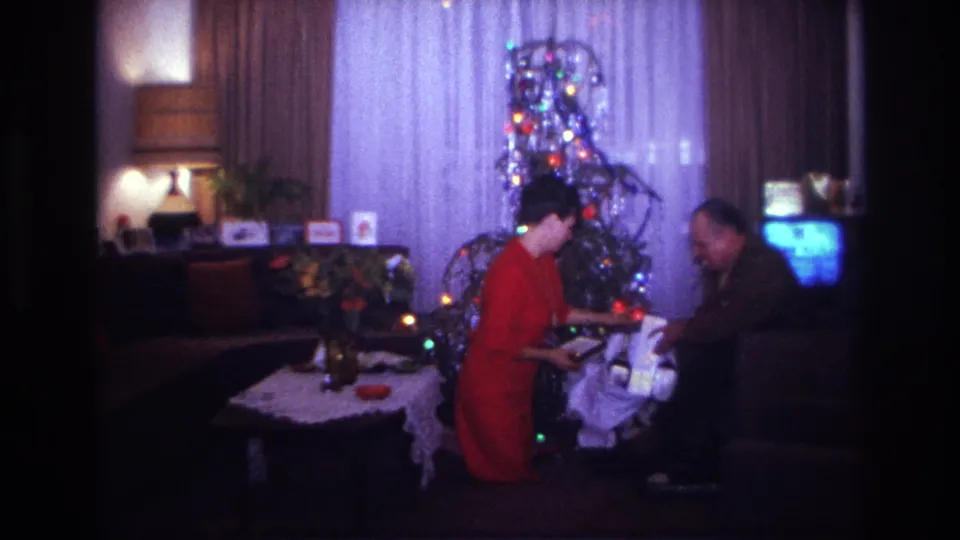 1967 Rockaway Beach home-movie frame showing adults sorting Christmas gifts in a private living room.