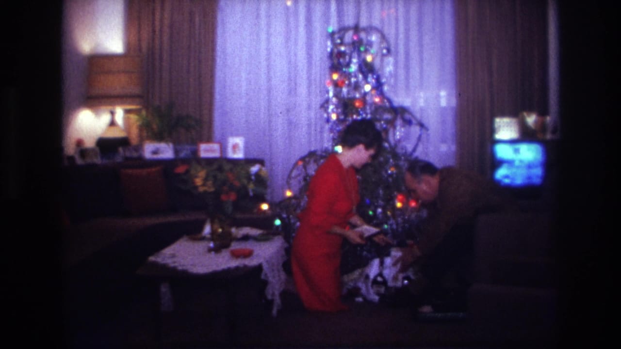 1967 Rockaway Beach home-movie frame showing two adults arranging gifts near a tinsel Christmas tree in a private living room.