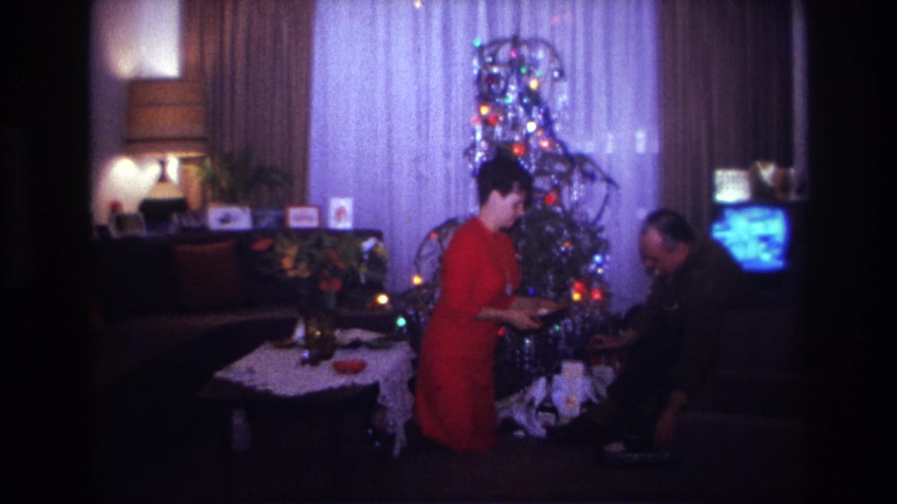 1967 Rockaway Beach home-movie frame with a woman in red and a man arranging gifts beside a lit tinsel Christmas tree.