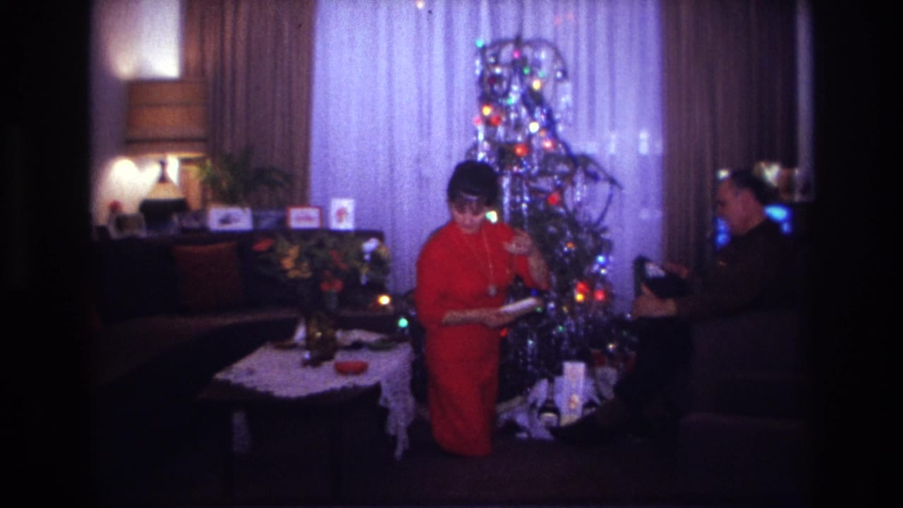 1967 Rockaway Beach home-movie frame showing a woman in a red dress decorating a lit Christmas tree as a man watches in a living room.